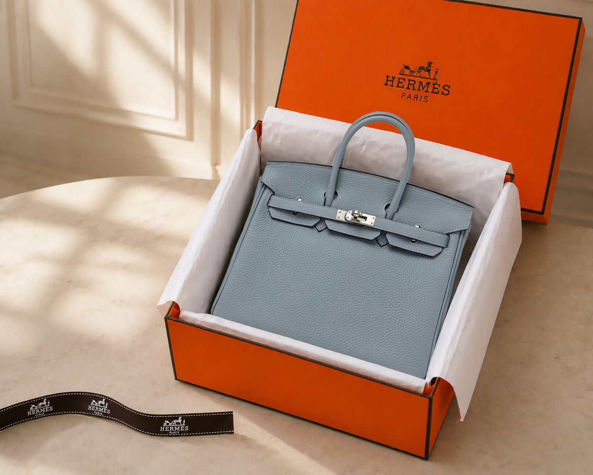Gray structured handbag with top handles and silver clasp inside an open orange Hermes box