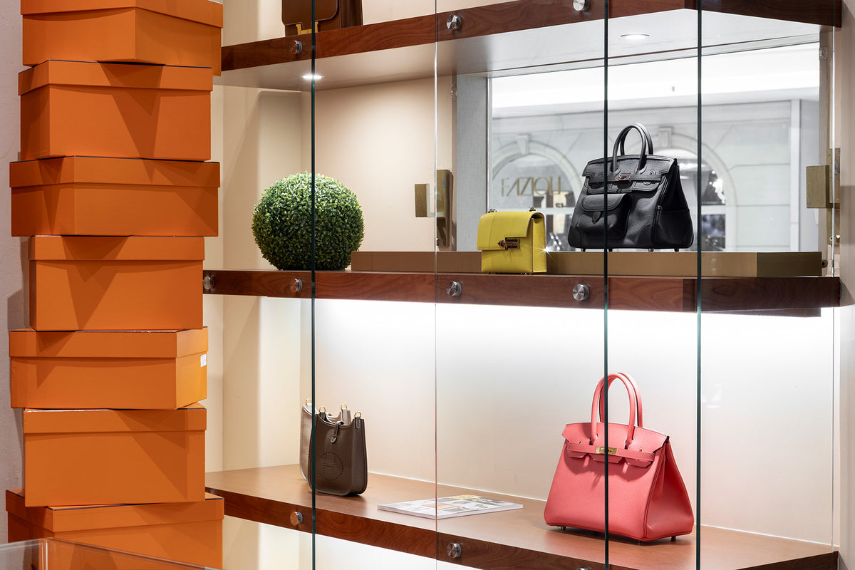 Glass display cabinet with black yellow pink and brown handbags beside stacked orange boxes
