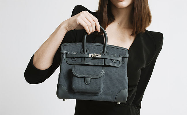 A woman in a black outfit holds up a structured handbag with top handles and a front flap.