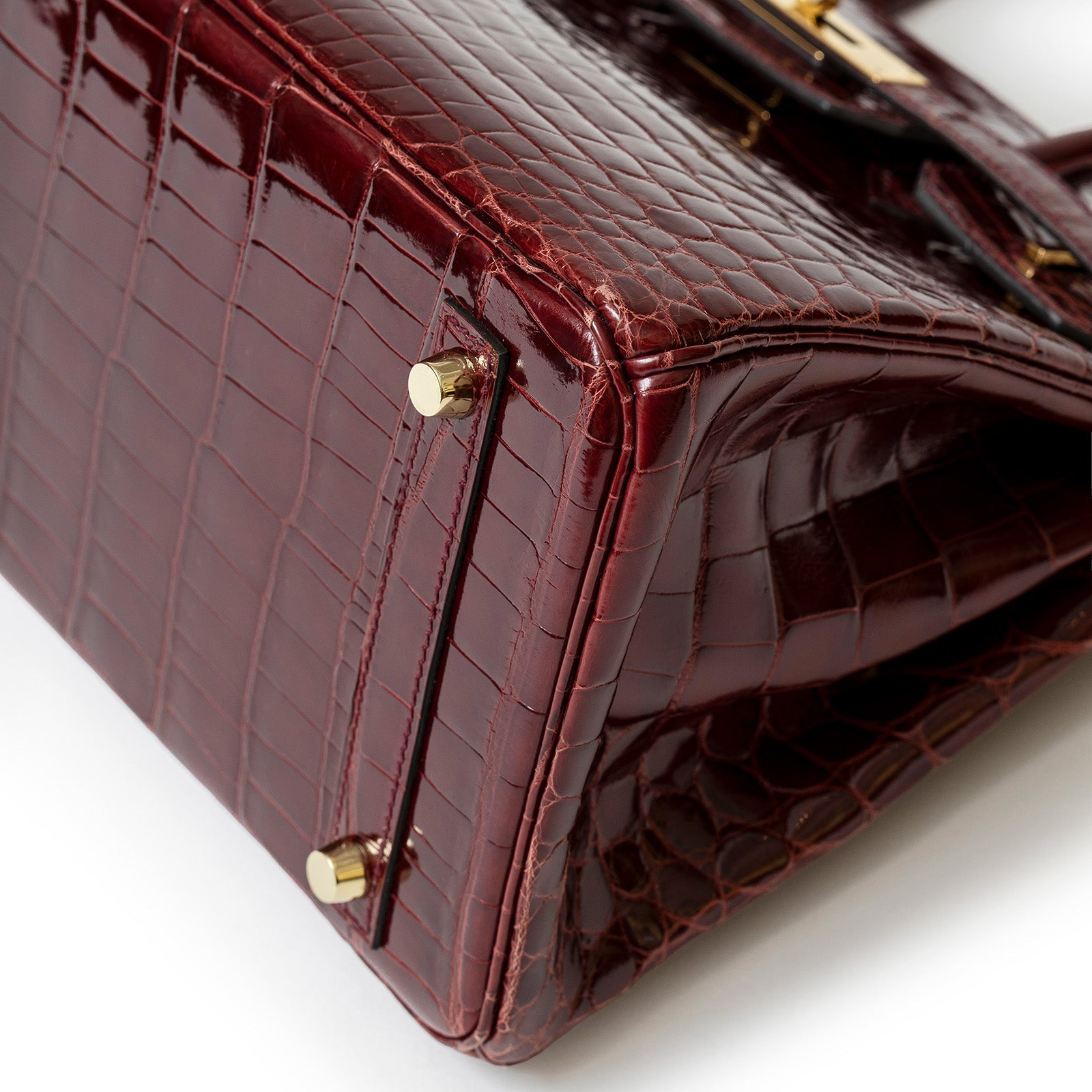 Close-up of the bottom corner showing shiny bourgogne crocodile texture and gold hardware feet on bag base
