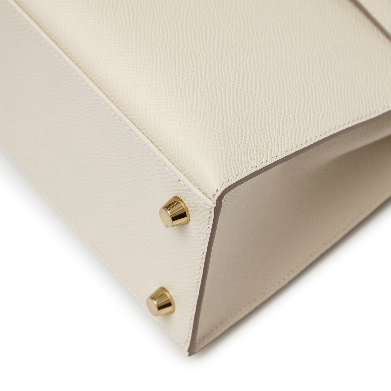 Close-up angled view of bottom corner of Hermès Kelly 25 Sellier Epsom Craie Permabrass Hardware handbag showing gold feet and textured leather