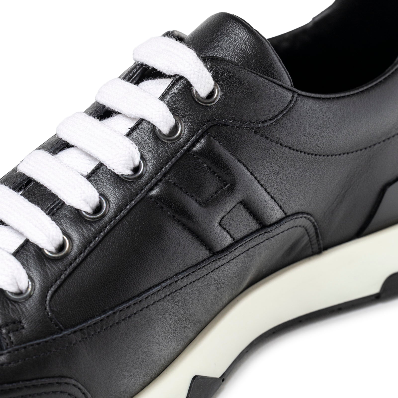 Close-up angled view of Hermès Men's Trail Sneaker Black showcasing leather, embossed logo, and white laces detail