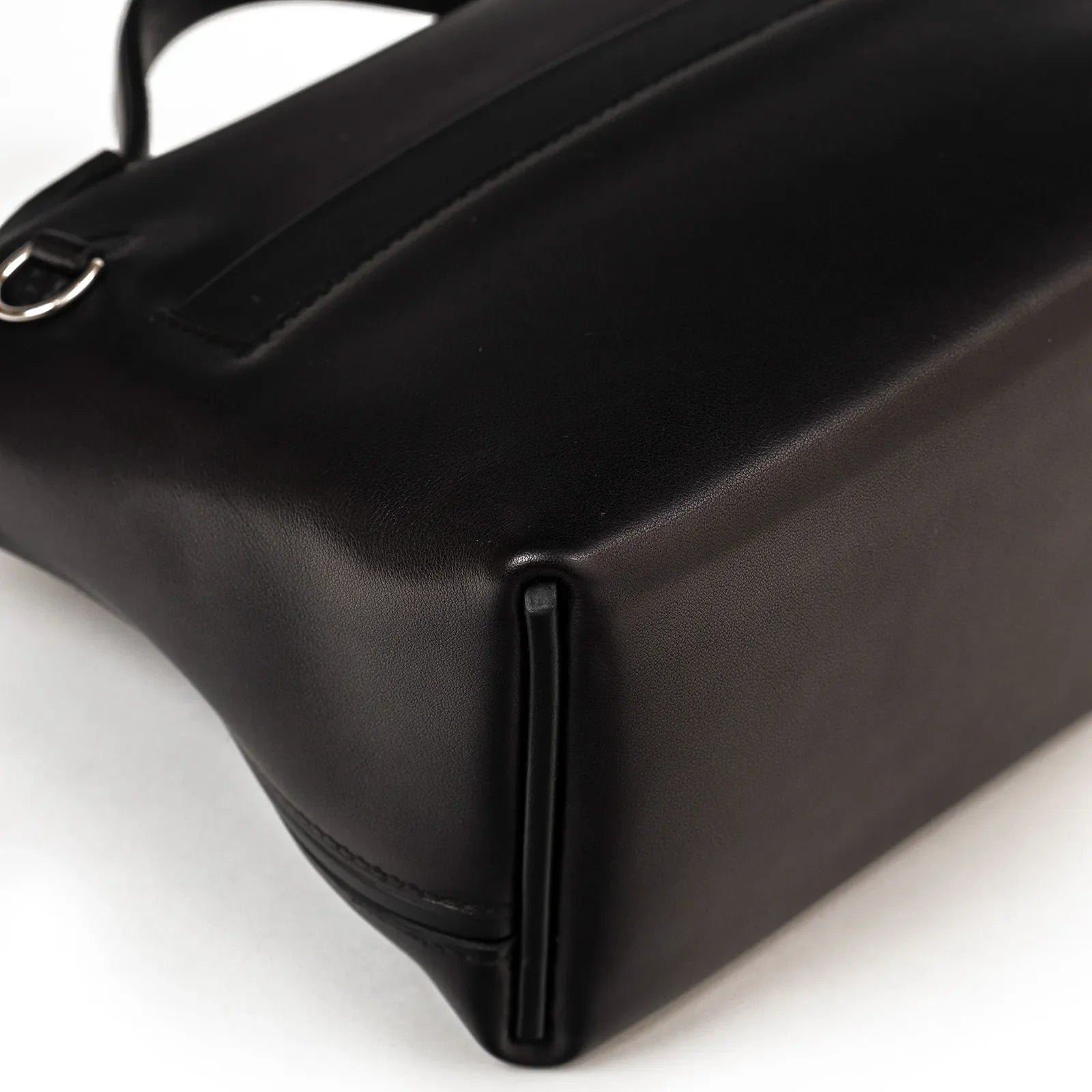 Close-up angled view of smooth black leather corner and palladium hardware on luxury handbag bottom edge.