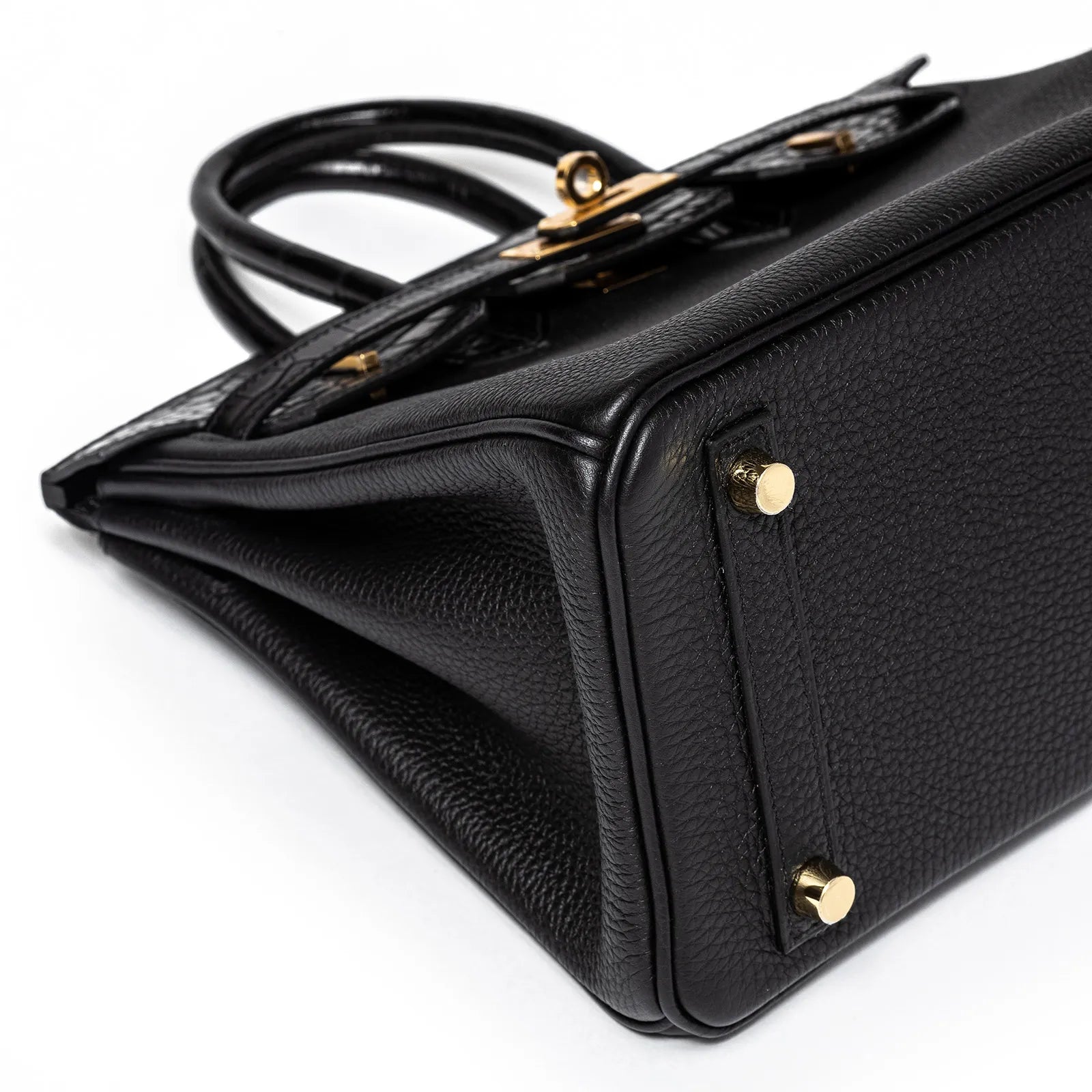 Close-up angled view of black togo leather base with gold hardware feet and matte alligator trim on bag edge
